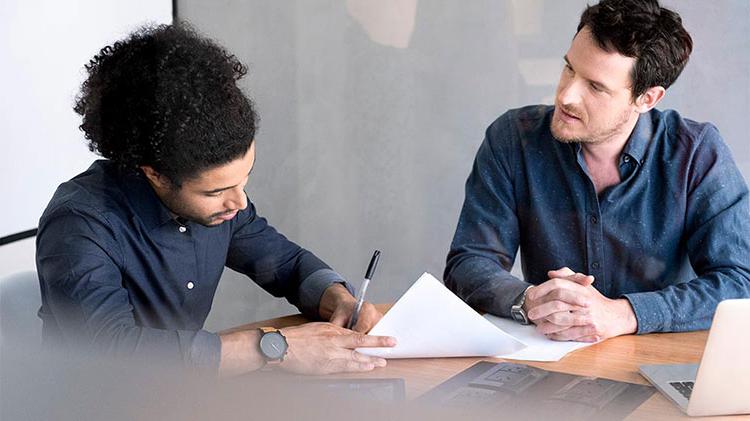 Two men sitting at a table and one of them is signing a car loan.